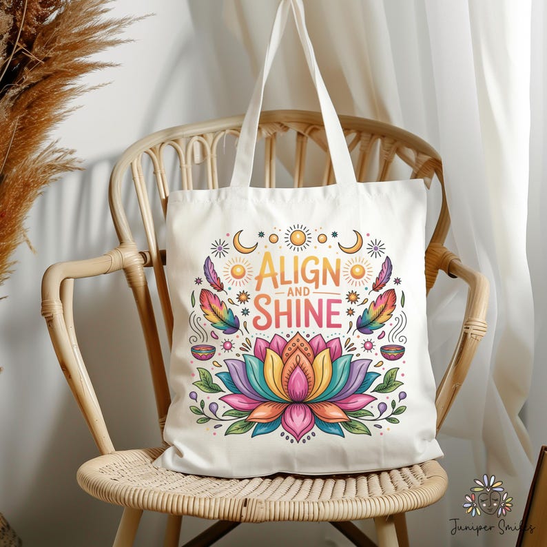 K&ouml;nnte beinhalten: Cremefarbene Tragetasche mit dem Schriftzug "ALIGN and SHINE" in einem farbenfrohen Design. Das Design umfasst eine Lotusblume, Federn, Sonnen, Monde und Sterne. Die Tasche liegt auf einem Korbstuhl.