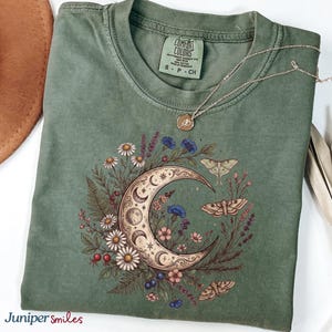 May include: A sage green t-shirt with a crescent moon and floral design. The moon graphic features a detailed celestial design with stars, flowers, and moths. A silver necklace with a small pendant rests on the shirt.