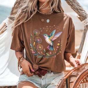 Op de afbeelding: Bruin T-shirt met een kleurrijk kolibrie- en bloemendesign. De illustratie toont een kolibrie met kleurrijke vleugels, omringd door paardenbloemen en andere bloemen. Het T-shirt heeft een relaxte pasvorm en is gemaakt van zacht materiaal.