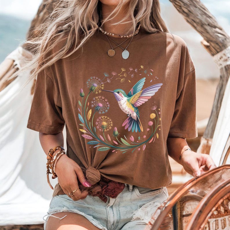 Op de afbeelding: Bruin T-shirt met een kleurrijk kolibrie- en bloemendesign. De illustratie toont een kolibrie met kleurrijke vleugels, omringd door paardenbloemen en andere bloemen. Het T-shirt heeft een relaxte pasvorm en is gemaakt van zacht materiaal.