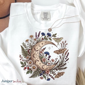 May include: White sweatshirt with a celestial design featuring a crescent moon, flowers, and moths. The design includes daisies, blue cornflowers, and a detailed moon phase illustration. A delicate necklace with a pendant rests on the sweatshirt. The brand name "Comfort Colors" is visible.
