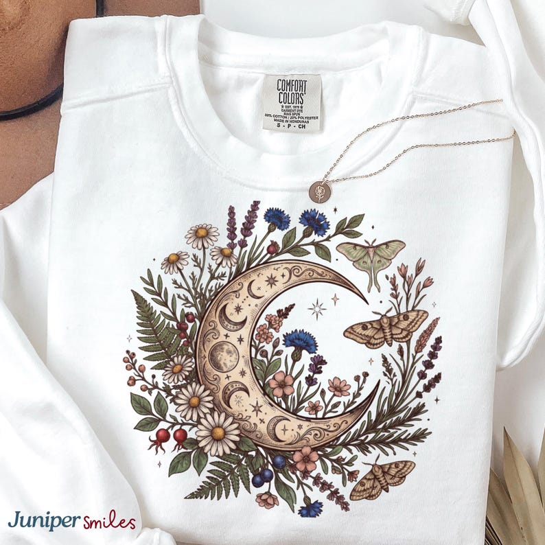 May include: White sweatshirt with a celestial design featuring a crescent moon, flowers, and moths. The design includes daisies, blue cornflowers, and a detailed moon phase illustration. A delicate necklace with a pendant rests on the sweatshirt. The brand name "Comfort Colors" is visible.