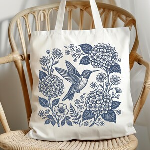 May include: White tote bag featuring a blue hummingbird design surrounded by flowers and foliage. The illustration includes hydrangeas, daisies, and other floral elements. The bag has two white handles and is sitting on a wicker chair.