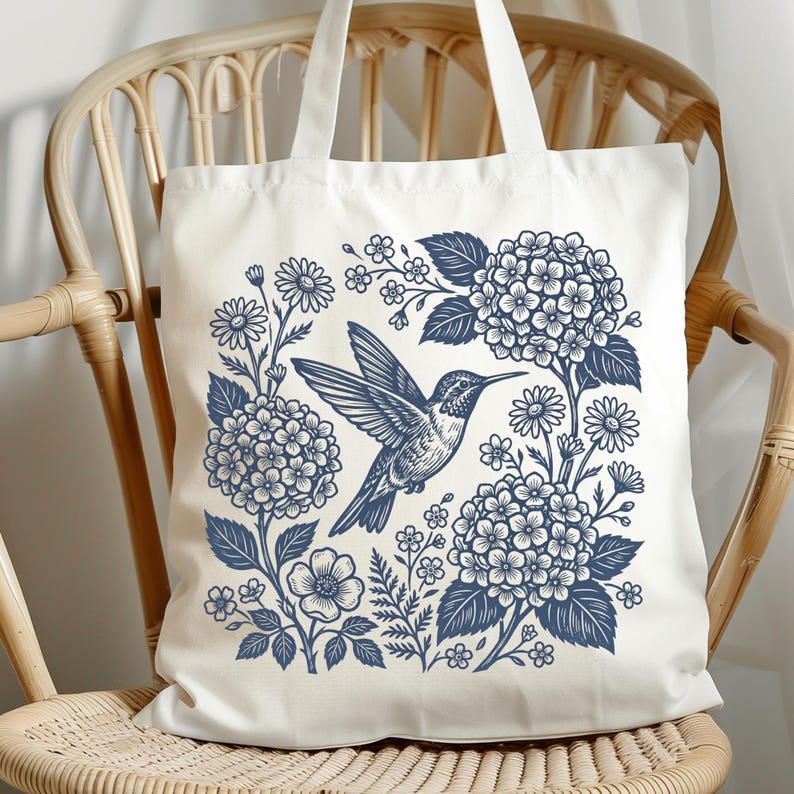 May include: White tote bag featuring a blue hummingbird design surrounded by flowers and foliage. The illustration includes hydrangeas, daisies, and other floral elements. The bag has two white handles and is sitting on a wicker chair.