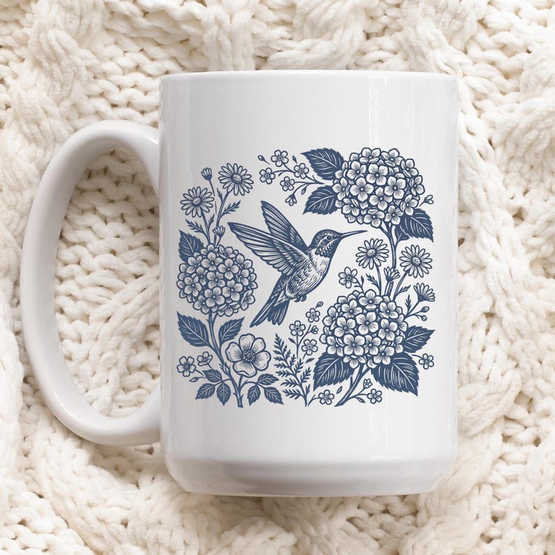 May include: White ceramic mug featuring a blue hummingbird design surrounded by flowers and foliage. The mug has a large handle and a straight, cylindrical shape. The floral design includes hydrangeas and daisies.