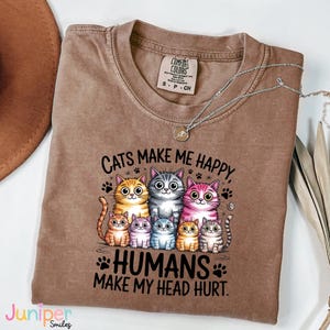 Puede incluir: Camiseta Comfort Colors marr&oacute;n con el texto "Cats Make Me Happy, Humans Make My Head Hurt". La camiseta presenta un gr&aacute;fico de varios gatos de dibujos animados en varios colores. Un collar plateado con un colgante est&aacute; sobre la camiseta.