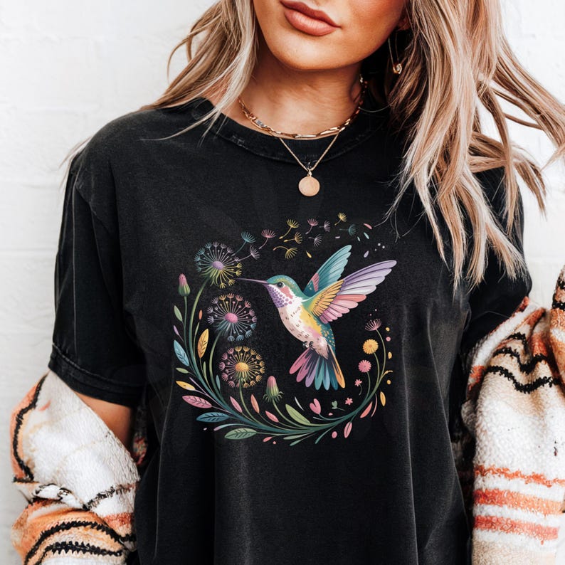 Op de afbeelding: Zwart T-shirt met een kleurrijk kolibrie-ontwerp. De illustratie toont een kolibrie met heldere vleugels, omringd door paardenbloemen en bloemelementen. Het shirt is donker van kleur, met een licht vervaagde uitstraling.