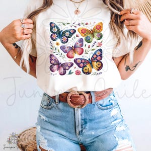 May include: A cream-coloured t-shirt displaying a vibrant butterfly design. The butterflies are decorated with floral patterns in blue, yellow, pink, and purple. The shirt is worn with blue denim shorts and a brown belt with a decorative buckle.
