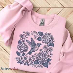 May include: A light pink sweatshirt featuring a detailed illustration of a hummingbird surrounded by flowers and foliage in shades of blue. The text "Juniper smiles" is at the bottom left. The sweatshirt is made of a soft material.