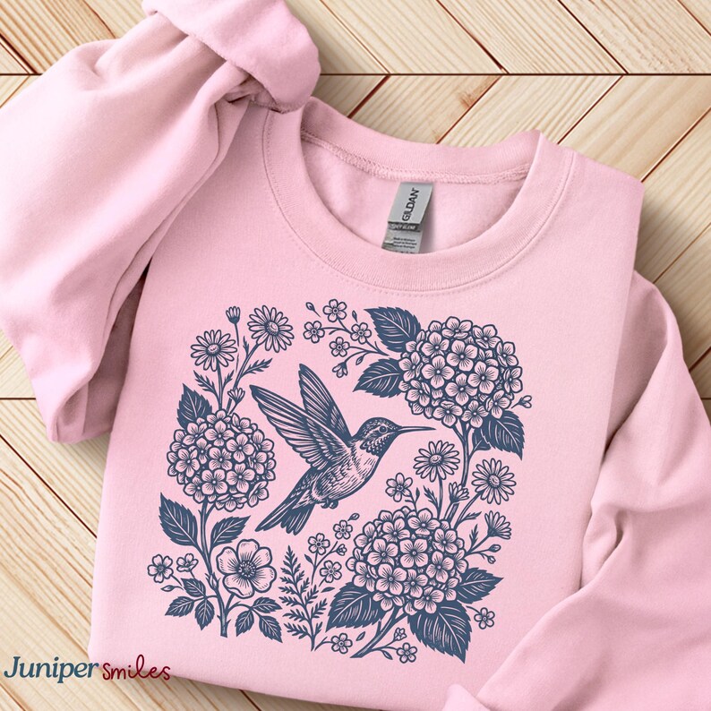 May include: A light pink sweatshirt featuring a detailed illustration of a hummingbird surrounded by flowers and foliage in shades of blue. The text "Juniper smiles" is at the bottom left. The sweatshirt is made of a soft material.