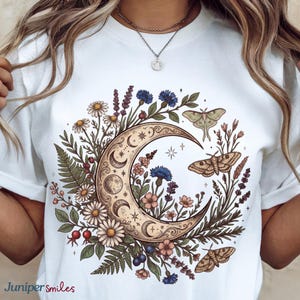 May include: White t-shirt featuring a crescent moon design with floral and moth illustrations. The design includes daisies, lavender, and blue flowers, with a detailed, vintage aesthetic. The text "Juniper smiles" is at the bottom left.