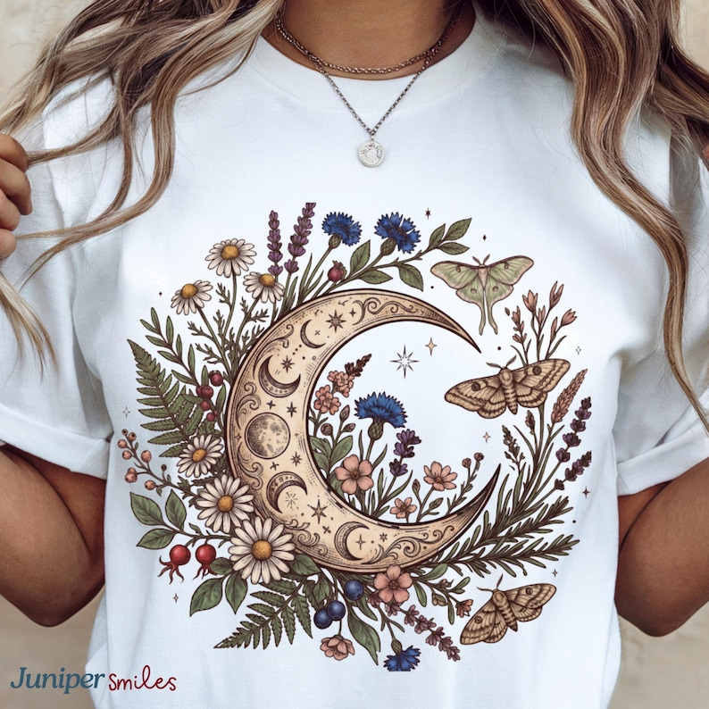 May include: White t-shirt featuring a crescent moon design with floral and moth illustrations. The design includes daisies, lavender, and blue flowers, with a detailed, vintage aesthetic. The text "Juniper smiles" is at the bottom left.