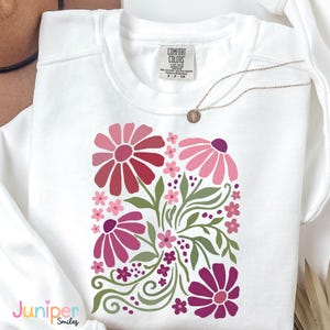 May include: White sweatshirt with a floral design in shades of pink and red, with green leaves and stems. The sweatshirt has a crew neck and long sleeves. The brand label "Comfort Colors" is visible. A gold necklace with a pendant is also present.
