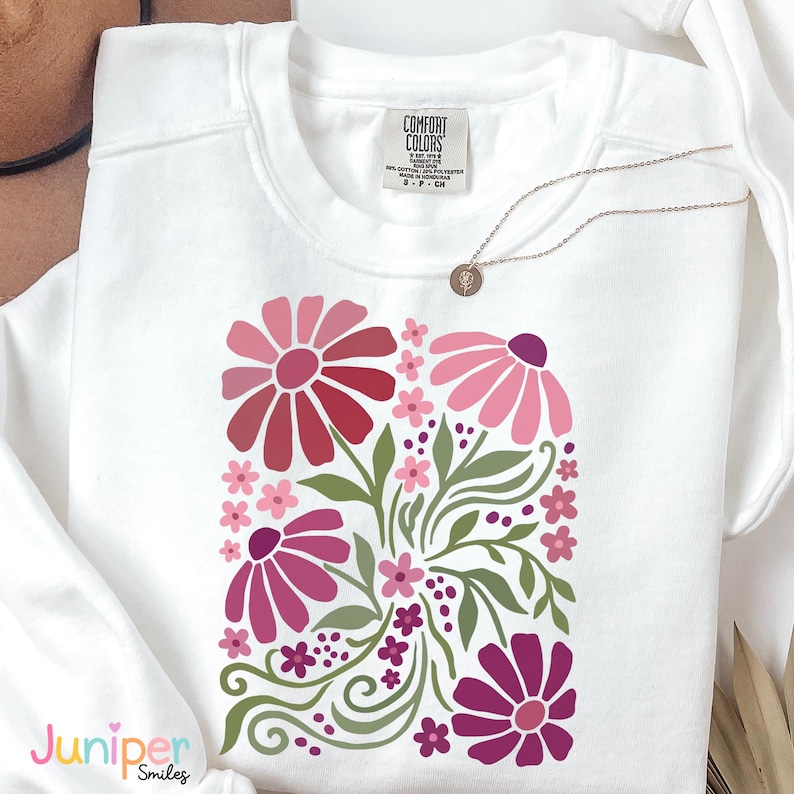 May include: White sweatshirt with a floral design in shades of pink and red, with green leaves and stems. The sweatshirt has a crew neck and long sleeves. The brand label "Comfort Colors" is visible. A gold necklace with a pendant is also present.