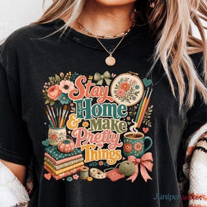 May include: A black t-shirt featuring a colorful graphic with the text Stay Home & Make Pretty Things. The design includes illustrations of paintbrushes, an embroidery hoop, a mug, stacked fabric, a pincushion, yarn, and a measuring tape.