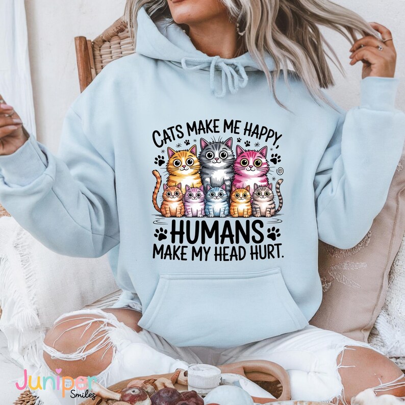 Puede incluir: Sudadera con capucha azul claro con un gr&aacute;fico de siete gatos de dibujos animados de varios colores. El texto dice: "Los gatos me hacen feliz, los humanos me dan dolor de cabeza". El dise&ntilde;o incluye huellas de patas y estrellas.