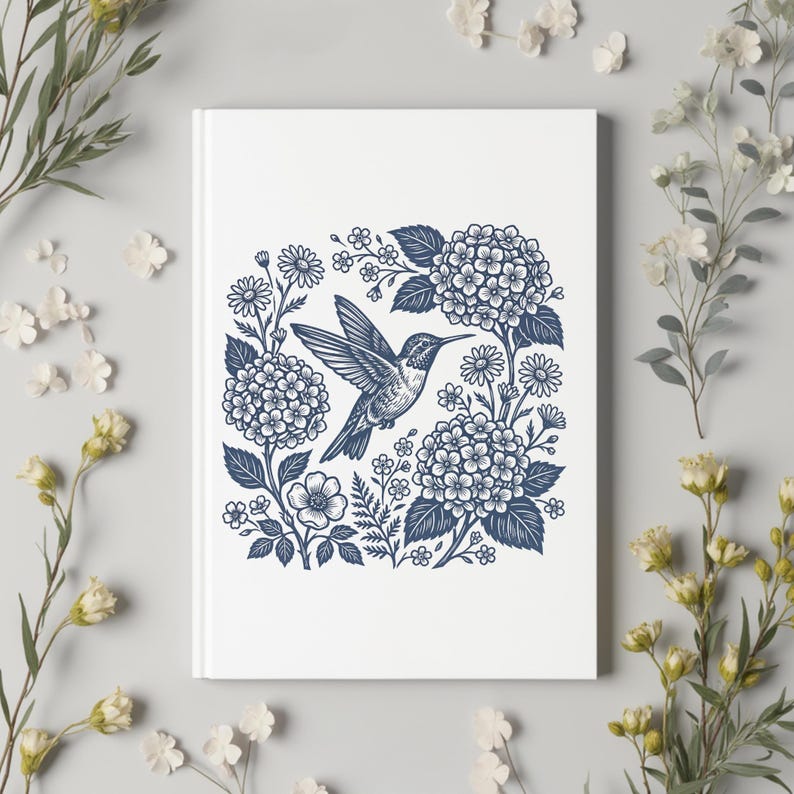 May include: White journal with a blue and white illustration of a hummingbird surrounded by flowers. The design features hydrangeas, daisies, and other floral elements. The cover is clean and simple, with a detailed nature-themed graphic.