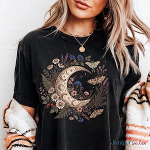 May include: Black t-shirt featuring a crescent moon design with floral and moth illustrations. The moon is surrounded by daisies, ferns, and other flowers. The shirt has a vintage, worn look.