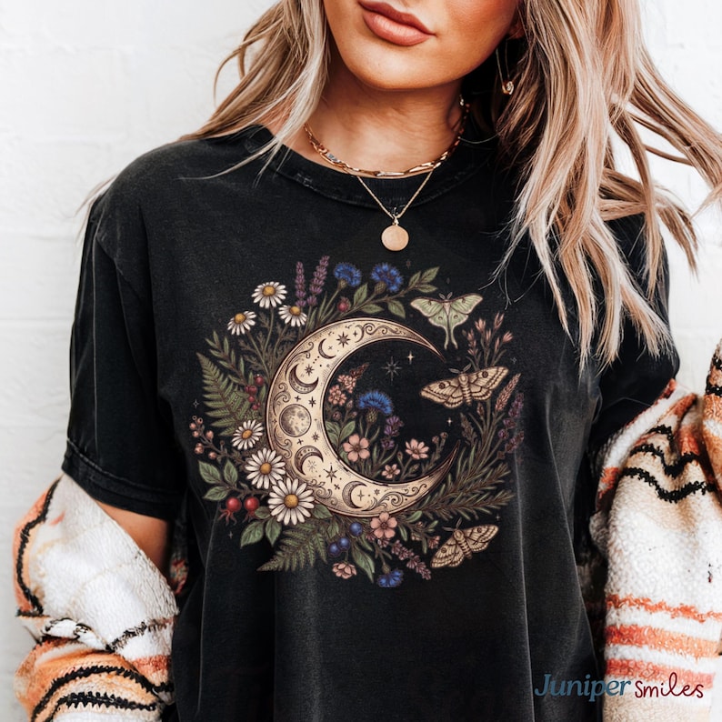 May include: Black t-shirt featuring a crescent moon design with floral and moth illustrations. The moon is surrounded by daisies, ferns, and other flowers. The shirt has a vintage, worn look.