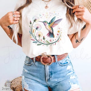 Könnte beinhalten: Cremefarbenes T-Shirt mit einem farbenfrohen Kolibri-Design in einem Blumenkranz. Der Kolibri hat blaue, grüne und rosa Federn. Das Shirt wird mit Jeansshorts und einem braunen Gürtel mit dekorativer Schnalle getragen.