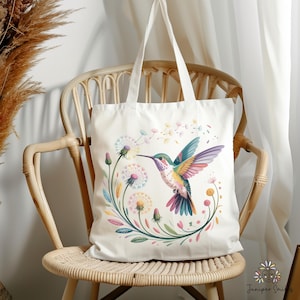 Op de afbeelding: Cr&egrave;mekleurige draagtas met een kleurrijk kolibrie-ontwerp. De vogel heeft heldere vleugels en een lange snavel, omringd door paardenbloemen en bloemmotieven. De tas heeft lange handvatten en staat op een rotan stoel.