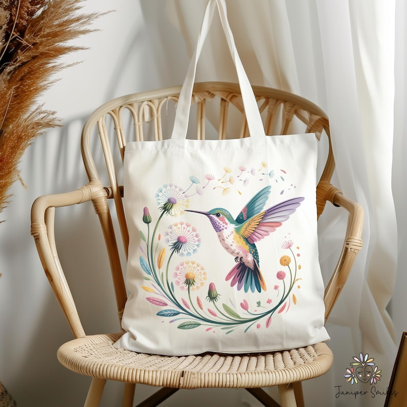 Op de afbeelding: Cr&egrave;mekleurige draagtas met een kleurrijk kolibrie-ontwerp. De vogel heeft heldere vleugels en een lange snavel, omringd door paardenbloemen en bloemmotieven. De tas heeft lange handvatten en staat op een rotan stoel.