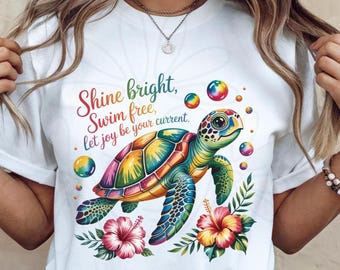 Pode incluir: Uma t-shirt branca com uma ilustração colorida de uma tartaruga marinha, flores de hibisco cor-de-rosa e amarelas e bolhas flutuantes. O texto diz: Shine bright, Swim free, let joy be your current. O casco da tartaruga exibe um gradiente arco-íris.