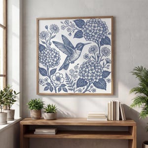 May include: A framed print featuring a blue hummingbird in flight, surrounded by detailed illustrations of hydrangeas, daisies, and other floral elements. The artwork is set against a white background, framed in a natural wood tone, and displayed on a wooden table.
