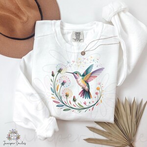 Op de afbeelding: Witte sweatshirt met een kleurrijk kolibrie-ontwerp en een bloemenkrans. De kolibrie heeft levendige vleugels en een lange snavel, omringd door paardenbloemen en bladeren. Een gouden ketting en een bruine hoed zijn ook zichtbaar.