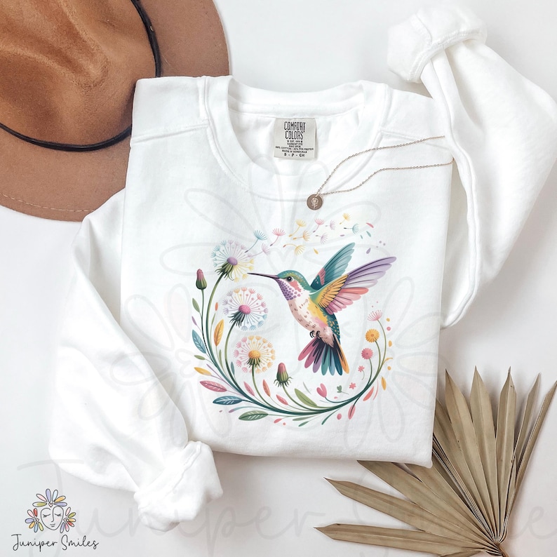 Op de afbeelding: Witte sweatshirt met een kleurrijk kolibrie-ontwerp en een bloemenkrans. De kolibrie heeft levendige vleugels en een lange snavel, omringd door paardenbloemen en bladeren. Een gouden ketting en een bruine hoed zijn ook zichtbaar.