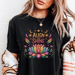 K&ouml;nnte beinhalten: Schwarzes T-Shirt mit einem farbenfrohen Lotusblumen-Design und dem Text "Align and Shine". Die Grafik enth&auml;lt Monde, Sonnen und Federn. Das Shirt ist ein bequemer, l&auml;ssiger Stil.