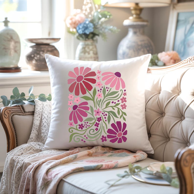 May include: White square throw pillow with a floral design in shades of pink and red. The flowers are arranged in a vertical pattern with green leaves and stems. The pillow is on a beige tufted sofa.