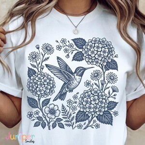 May include: White t-shirt featuring a detailed blue illustration of a hummingbird in flight, surrounded by flowers and foliage. The design includes hydrangeas, daisies, and other floral elements. The text "Juniper Smiles" is at the bottom.