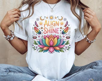 May include: White t-shirt featuring a colorful floral design with the text "ALIGN and SHINE". The design includes a lotus flower, feathers, moons, and suns. The shirt is a comfortable, casual style.