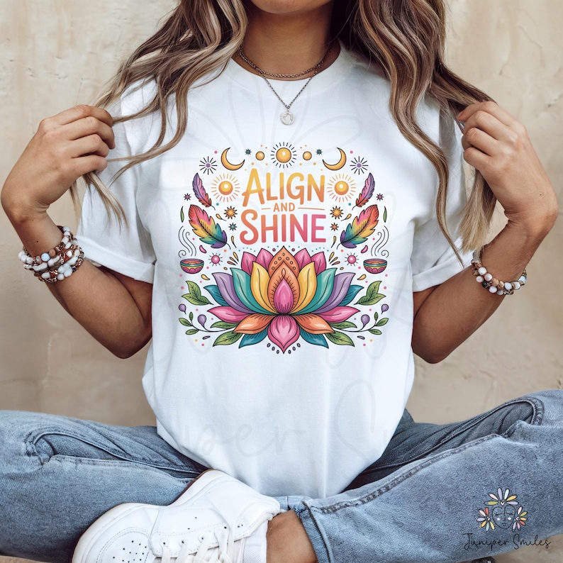 K&ouml;nnte beinhalten: Wei&szlig;es T-Shirt mit einem farbenfrohen Blumendesign und dem Text "ALIGN and SHINE". Das Design beinhaltet eine Lotusblume, Federn, Monde und Sonnen. Das T-Shirt ist ein l&auml;ssiger Stil.