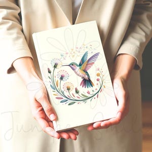 Op de afbeelding: Een cr&egrave;mekleurig notitieboek met een kleurrijk kolibrie-ontwerp. De kolibrie is omgeven door een bloemenkrans met paardenbloemen en andere bloemen. Het notitieboek wordt voor een neutrale achtergrond gehouden.