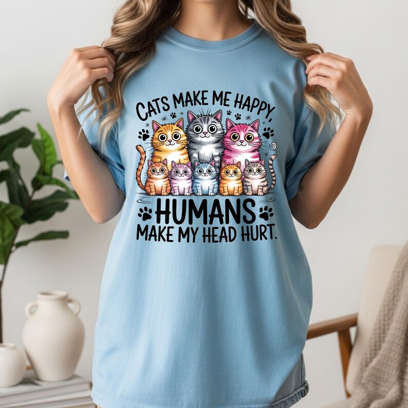 Puede incluir: Camiseta azul claro con un gr&aacute;fico de siete gatos de dibujos animados de varios colores. El texto "CATS MAKE ME HAPPY, HUMANS MAKE MY HEAD HURT" est&aacute; impreso encima y debajo de los gatos. Huellas de patas dispersas.