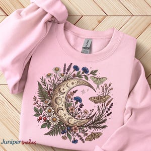 May include: A light pink sweatshirt featuring a crescent moon design with floral and moth illustrations. The moon is surrounded by daisies, blue flowers, and ferns. The text "Juniper smiles" is in the bottom left corner.