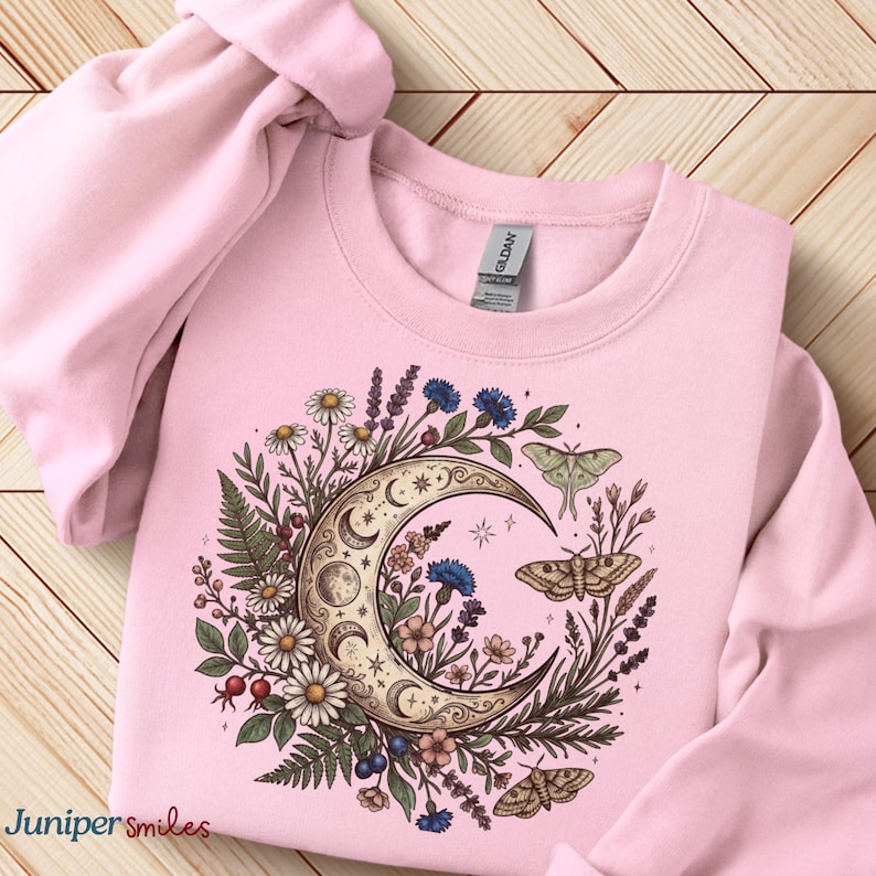 May include: A light pink sweatshirt featuring a crescent moon design with floral and moth illustrations. The moon is surrounded by daisies, blue flowers, and ferns. The text "Juniper smiles" is in the bottom left corner.