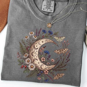 May include: Gray t-shirt with a crescent moon and floral design. The moon is surrounded by daisies, blue cornflowers, and moths. A silver necklace with a small pendant rests on the shirt. The tag reads "COMFORT COLORS".