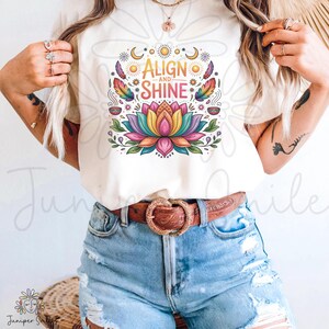 K&ouml;nnte beinhalten: Cremefarbenes T-Shirt mit einem farbenfrohen Lotusblumen-Design und dem Text "ALIGN and SHINE". Das Design beinhaltet Monde, Sonnen, Federn und Bl&auml;tter. Das Shirt wird mit blauen Jeansshorts und einem braunen G&uuml;rtel kombiniert.