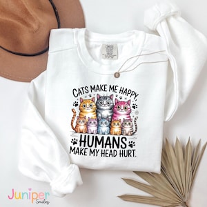 Puede incluir: Sudadera blanca con cuello redondo y un dise&ntilde;o de siete gatos de dibujos animados de varios colores. El texto dice: "Cats make me happy, humans make my head hurt."