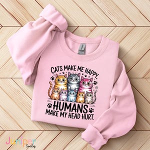 Puede incluir: Sudadera rosa claro con el texto "Cats make me happy, humans make my head hurt". El dise&ntilde;o presenta un grupo de gatos de dibujos animados en varios colores. La sudadera tiene mangas largas y cuello redondo.