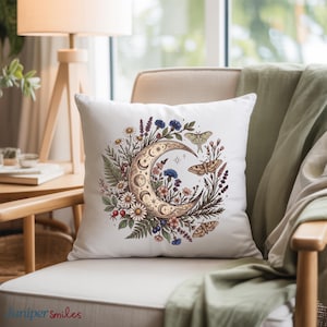 May include: White square throw pillow featuring a crescent moon design filled with flowers, leaves, and butterflies. The pillow has a botanical theme with a gold moon and colorful floral accents. The pillow is on a beige chair.