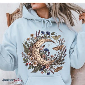 May include: Light blue hoodie featuring a crescent moon design with floral and moth illustrations. The moon is surrounded by daisies, blue cornflowers, and other wildflowers. The text "Juniper smiles" is at the bottom left.