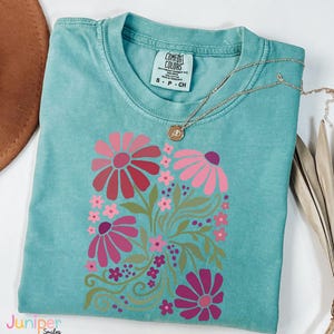 May include: A turquoise Comfort Colors t-shirt featuring a floral design in shades of pink and red. The design includes large and small flowers with green leaves and stems. A gold necklace with a pendant rests on the shirt.