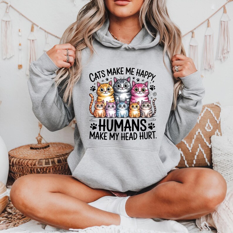 Puede incluir: Una sudadera con capucha gris claro con un gr&aacute;fico de seis gatos de dibujos animados de varios colores. El texto "Cats make me happy, humans make my head hurt" est&aacute; impreso encima y debajo de los gatos. La sudadera tiene una capucha con cord&oacute;n.