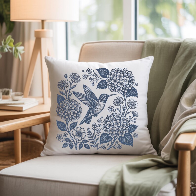 May include: Square white throw pillow featuring a blue hummingbird design surrounded by blue flowers and foliage. The pillow is placed on a beige chair with a green blanket draped over the arm. The design is a nature-themed decorative item.