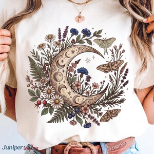 May include: Cream-colored t-shirt featuring a crescent moon design surrounded by flowers, leaves, and moths. The design includes daisies, blue cornflowers, and lavender. The text "Juniper smiles" is at the bottom left.