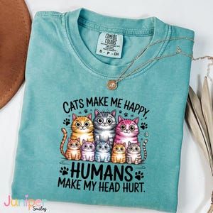 Puede incluir: Una camiseta turquesa Comfort Colors con el texto "Cats Make Me Happy, Humans Make My Head Hurt". La camiseta presenta un gr&aacute;fico de varios gatos de dibujos animados. Un collar dorado con un colgante est&aacute; drapeado sobre la camiseta.